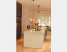 Moderne Küche mit einer großen Kücheninsel aus Beton, darüber zwei hängende Lampen. Im Hintergrund helle Vorhänge und große Fenster, die viel Licht in den Raum lassen, Holzboden im Fischgrätmuster.