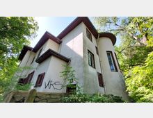 Wohnhaus mit heller Putzfassade, braunen Fensterläden und einem runden, turmartigen Bauelement, umgeben von hohen Bäumen und dichter Vegetation; im Vordergrund ein Holzzaun, auf der Wand steht ein Graffiti mit den Buchstaben „VRS“.