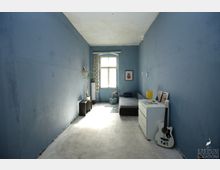 Ein schmaler Raum mit blauen Wänden, einem großen Fenster am Ende und weißem Boden. Die Einrichtung umfasst ein Sofa, eine weiße Kommode mit Bildern und einer Gitarre, sowie eine Topfpflanze und eine Stehlampe in einer Ecke.