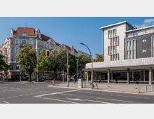 Straßenszene mit einer Kreuzung, flankiert von einem historischen Gebäude mit ornamentalen Fassaden und roten Dächern links sowie einem modernen, mehrstöckigen Gebäude mit geometrischer Fassade und bunten Fensterakzenten rechts; dazwischen Bäume und Verkehrsampeln.