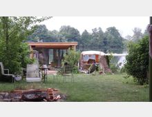 Ein Garten mit Feuerstelle im Vordergrund, verschiedenen Gartenmöbeln und Blick auf einen kleinen, modernen Holzpavillon am Ufer eines Sees. Im Hintergrund sind dichte, grüne Bäume zu sehen, die das Wasser umrahmen.