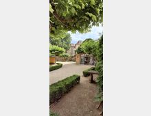 Innenhofgarten mit Kieswegen, niedrigen Buchshecken und dichtem Blätterdach aus Bäumen. Rechts steht ein kleiner Backsteinbau mit großer Glasfront, im Hintergrund eine Steinmauer und ein mehrstöckiges Wohnhaus.