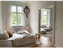 Schlafzimmer mit hellen Wänden, einem Bett mit weißer Bettwäsche und einem großen Fenster mit Blick auf grüne Bäume; daneben ein offener Durchgang zu einem Wohnzimmer mit weißem Sofa und einem Couchtisch. Holzboden und eine Hängelampe aus geflochtenem Material betonen die Einrichtung.