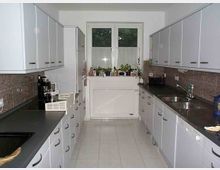 Helle Einbauküche mit zwei parallelen Arbeitsflächen, weißen Hängeschränken, grauen Arbeitsplatten und einem zentralen Fenster mit Topfpflanzen auf der Fensterbank. Die Rückwand ist mit braunen Fliesen versehen, und Elektrogeräte wie Kaffeemaschine und Toaster sind sichtbar.