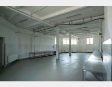 Großer, leerer Raum mit grauem Boden und weißen Wänden, teilweise mit Fliesen verkleidet. Der Raum hat eine Industrieküchen-ähnliche Einrichtung mit mehreren Wandtischen aus Edelstahl sowie sichtbaren Rohren und Deckeninstallationen.