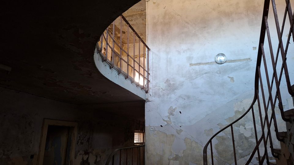 Innenansicht eines heruntergekommenen Treppenhauses in einem alten Gebäude mit abblätterndem Putz und rissigen Wänden. Rechts führt eine geschwungene Metalltreppe nach oben zu einem halb offenen Rundgang mit Geländer, beleuchtet durch ein kleines Fenster und eine einfache Wandlampe.