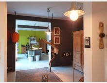 Wohnbereich mit offener Verbindung zur Küche im Hintergrund. Der Raum ist dekoriert mit Holzschränken, farbigen Wänden (darunter eine grüne Küchenwand), Hängelampen, Kunstwerken an der Wand und einem Teppich auf dem Boden; ein kleiner Hund steht in der Mitte.