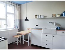 Helle Küche im schlichten Design mit weißen Schränken, einer großen Keramikspüle, einer Arbeitsplatte mit Holzakzent und einem kleinen Esstisch mit Hocker. Ein großes Fenster mit Vorhängen lässt viel Tageslicht herein; darüber hinaus gibt es eine schlichte schwarze Hängeleuchte.