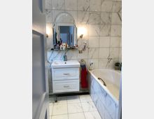 Badezimmer mit weißem Marmordesign an den Wänden und dem Boden. Es verfügt über ein weißes Waschbecken mit Unterschrank, darüber ein Spiegel mit geschwungenem Rahmen und eine Badewanne rechts davon.