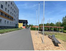 Ein moderner Außenbereich mit einer gepflasterten Gehwegfläche, Bänken und ungenutzten Fahnenmasten. Links steht ein mehrstöckiges Gebäude mit grauer und gelber Fassade, im Hintergrund sind eine Fahrradüberdachung sowie grüne Vegetation zu sehen.