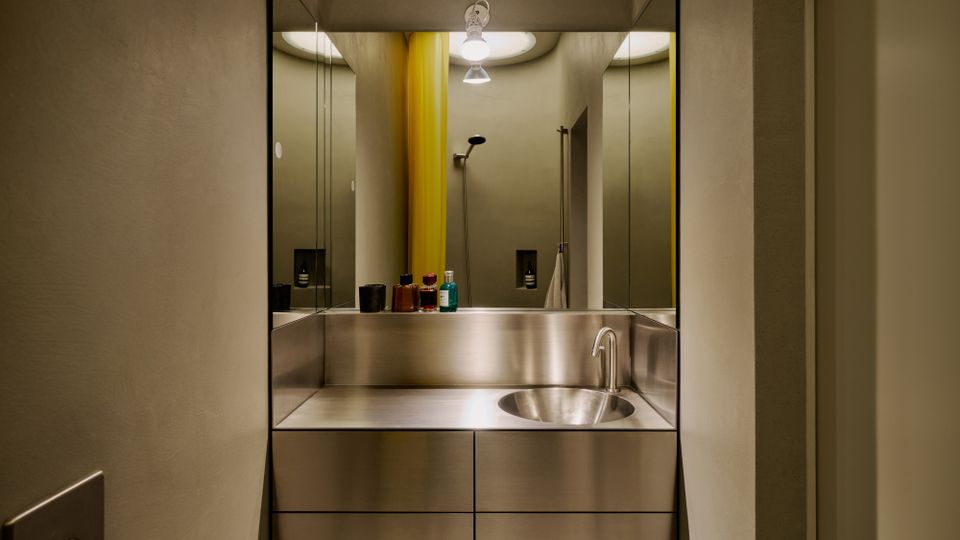 Modernes Badezimmer mit wandhängendem Waschtisch aus Edelstahl und rundem Waschbecken, davor eine große Spiegelwand. Im Hintergrund sind eine Dusche mit gelbem Duschvorhang, eingelassene Nischen und eine Deckenleuchte zu sehen.