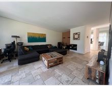 Wohnzimmer mit heller Fliesenbodenoptik, einer großen dunklen Eckcouch, einem bunten Holztisch und einer Wanddekoration aus einem Gemälde in Türkis und Gold sowie einem Kamin an einer hellen Trennwand. Im Hintergrund befinden sich ein Arbeitsbereich und ein Esstisch bei bodentiefen Fenstern mit Vorhängen.