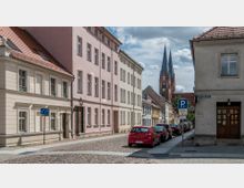 Pflastersteinstraße in einer Altstadt mit pastellfarbenen Gebäuden, parkenden Autos und der im Hintergrund sichtbaren roten Backsteinkirche mit hohen spitzen Kirchtürmen.