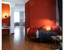 Ein Zimmer mit Holzboden und einer roten Wand, an der eine kleine Lampe befestigt ist. Im Vordergrund steht ein Bett mit bunten Kissen und Decken, daneben ein kleiner Tisch; im Hintergrund führt der Raum mit hellem Tageslicht und weißen Wänden in einen weiteren Bereich mit einem Fenster und Heizkörper.