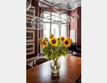 Ein stilvoller Innenraum mit einem Holztisch, auf dem eine Vase mit Sonnenblumen steht. Im Hintergrund befinden sich eine rote Wand mit gerahmten Bildern, ein Fenster mit Blick nach draußen und ein schwarzes Klavier. Über dem Tisch hängt eine moderne Hängeleuchte mit gläsernen Kugeln.