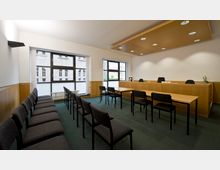 Ein moderner Gerichtssaal mit einer Reihe von grauen Sitzstühlen auf beiden Seiten, einem erhöhten Richtertisch aus Holz und einem einfachen Besprechungstisch mit Stühlen davor. Große Fenster lassen Tageslicht herein, während die Wand- und Deckenbeleuchtung den Raum gleichmäßig erhellt.