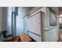 Innenansicht einer Kirche mit einer weißen Orgelverkleidung und sichtbaren silbernen Orgelpfeifen. Im Vordergrund stehen ein schlichter Holztisch und ein schwarzer Stuhl; im Hintergrund ist eine Säule sowie ein Fenster mit buntem Glas erkennbar.