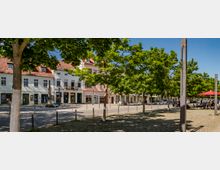 Straßenansicht eines Platzes mit Pflastersteinen, flankiert von Bäumen und Gebäuden mit rotem Ziegeldach und pastellfarbenen Fassaden. Im Vordergrund stehen vereinzelt Poller, während auf der rechten Seite Tische mit roten Sonnenschirmen unter freiem Himmel stehen.