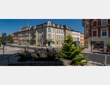 Kreuzung mit historischen Wohnhäusern, die durch verzierte Fassaden und bunte Farben geprägt sind, sowie Straßenschildern, einer Ampel und einem kleinen bepflanzten Bereich im Vordergrund. Die Gebäude haben mehrere Stockwerke und Dachgauben.