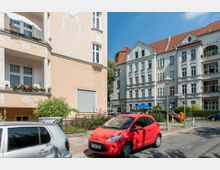 Straßenszene in einem Wohngebiet mit Altbauhäusern, die Fassaden mit weißen und beigefarbenen Putzdetails und Balkonen haben. Im Vordergrund parken ein rotes und ein silbernes Auto, daneben sind ein kleiner Garten und Straßenlaternen zu sehen.