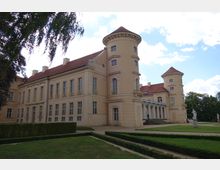 Historisches Schloss mit cremefarbener Fassade, roten Ziegeldächern und zwei runden Türmen. Im Vordergrund gepflegte Gartenanlagen mit Hecken und Rasenflächen, im Hintergrund blauer Himmel mit leichten Wolken.