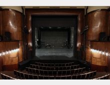 Blick in einen klassischen Theatersaal mit dunkler Bühne, hölzernen Wänden und einer Bestuhlung in mehreren Reihen. Die Bühne ist leer, und an den Seiten sind Licht- und Tonanlagen zu erkennen.