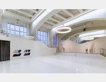 Großer, moderner Veranstaltungsraum mit hellen Wänden, glänzendem weißen Boden und hohen Decken mit dekorativen Details. Der Raum beinhaltet große, bogenförmige Fenster, eine erhöhte Bühne und eine runde Hängelampe als zentrales Designmerkmal.