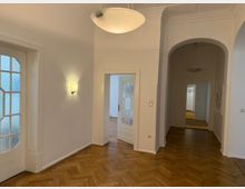 Heller Flur in einer Altbauwohnung mit Fischgrätparkett, hohen Decken, und weißen Wänden. Zu sehen sind zwei Türen mit Glaseinsätzen und ein abgerundeter Durchgang, der zu weiteren Räumen führt; an der Decke und Wänden befinden sich moderne Lampen.