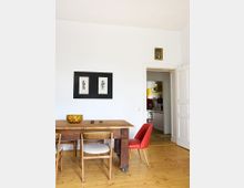 Essbereich mit einem Holztisch, umgeben von verschiedenen Stühlen, darunter ein roter Polsterstuhl. An der weißen Wand hängen zwei gerahmte Schwarz-Weiß-Zeichnungen und ein kleines Bild über einer Tür, die in eine Küche mit gelben Wandelementen führt.