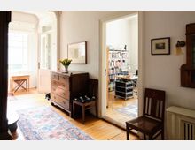 Ein heller Innenraum mit Holzboden, einem antiken Möbelstück mit Schubladen und einer Vase mit gelben Blumen darauf. Im Vordergrund führt eine Tür in einen angrenzenden Raum mit Bücherregalen, einem Bürotisch und Schreibtischlampe.