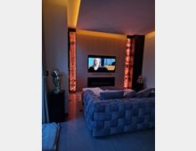 Modernes Wohnzimmer mit einem großen grauen Sofa im Vordergrund, einem an der Wand montierten Fernseher und beleuchteten Dekorelementen in Nischenregalen auf beiden Seiten. Die Beleuchtung ist warm und indirekt, mit dunklen Wänden und stilvollen goldenen Dekorobjekten.