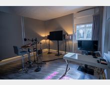 Ein modern eingerichteter Ton- oder Aufnahmebereich in einem Raum mit grauen Wänden und Holzboden. Sichtbar sind ein Mikrofon und Mischpult auf einem hohen Schreibtisch, ein großer Bildschirm mit Lautsprechern auf Stativen, ein kleiner Schreibtisch mit Monitor sowie ein Fenster mit Vorhängen, durch das Tageslicht einfällt.