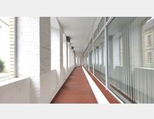 Ein überdachter Korridor mit weißen Backsteinwänden auf der linken Seite und großen Glasfenstern mit Jalousien auf der rechten Seite. Der Boden besteht aus roten Fliesen, und die Decke ist weiß gestrichen.