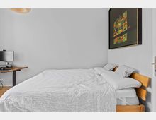 Schlafzimmer mit einem großen Bett mit weißer Bettwäsche und Holzrahmen, daneben ein höhenverstellbarer Schreibtisch mit Monitor. An der weißen Wand hängt ein farbenfrohes, rechteckiges Gemälde.