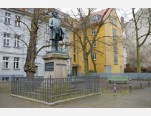 Statue auf einem kleinen Platz mit gepflastertem Boden, umgeben von kahlen Bäumen und einem niedrigen Metallzaun. Im Hintergrund stehen Wohnhäuser mit weißer und gelber Fassade.