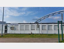 Ein eingeschossiges, weißes Containergebäude mit geschlossenen Rollläden, beschriftet mit "HLB-Hafenmeister", steht auf einem gepflasterten Gelände mit Rasenflächen. Im Hintergrund ist ein Brückengerüst sowie Stromleitungen sichtbar, darüber ein teils bewölkter Himmel.