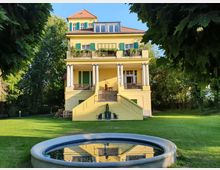Gelbes zweistöckiges Haus mit Balkon und grünen Fensterläden, im klassischen Stil mit Säulen und einem roten Ziegeldach. Im Vordergrund ein runder Brunnen, eingebettet in eine gepflegte Rasenfläche, umgeben von Bäumen.