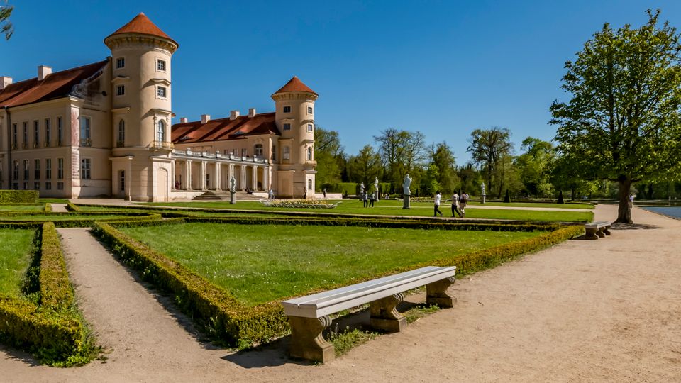 Large historic palace with cream-colored walls and red roofs, featuring two round corner towers, viewed across formal hedged garden paths. A sandy walkway runs along a lake on the right, lined with trees and a bench in the foreground.