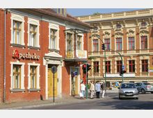 Straßenszene mit einem roten Backsteingebäude im Vordergrund, das eine Apotheke beherbergt, und einem dekorativen Sandsteingebäude mit aufwendiger Fassade im Hintergrund. Fußgänger stehen an einer Ampel, während ein Auto an der Kreuzung hält.