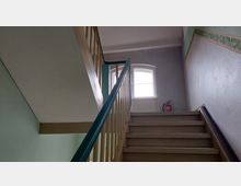 Treppenhaus mit cremefarbenen Stufen und einem grünen Geländer, hell erleuchtet durch ein großes, bogenförmiges Fenster im Hintergrund; neben den Stufen steht ein roter Feuerlöscher an der Wand.