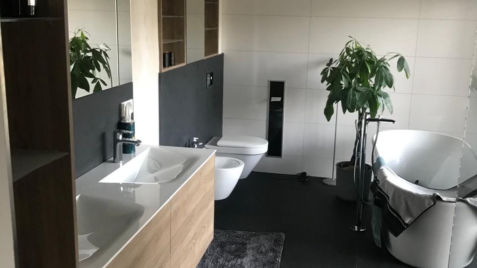 Modernes Badezimmer mit großformatigen hellen Wandfliesen und dunklen Bodenfliesen, Holz-Waschtisch mit Doppelwaschbecken und großem Spiegelschrank. Rechts stehen eine freistehende Badewanne mit Handtuch, ein bodenstehender Wasserhahn und eine große Topfpflanze.