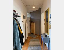 Schmaler Flur mit Holzdielenboden, links eine Garderobe mit Jacken, rechts ein kleiner Konsolentisch mit Schubladen und ein Spiegel an der Wand. Im Hintergrund steht eine schlichte Holzkommode vor einer grauen Wand, beleuchtet von einer einfachen Hängelampe.