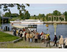 Eine Gruppe von Menschen steht an einer Anlegestelle und steigt auf ein kleines Ausflugsschiff mit gelbem Dach ein. Im Hintergrund ist ein breiter Fluss mit einem größeren weißen Passagierschiff, grüner Ufervegetation und einem Säulengang auf der gegenüberliegenden Seite zu sehen.