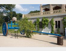 Ein dekorativer historischer blaulackierter zweirädriger Wagen steht auf einem Kiesplatz vor einer mit Rankpflanzen bedeckten Steinmauer. Im Hintergrund sind ein Balkon mit schmiedeeisernem Geländer und ein weißes Zierguss-Sitzmöbel sichtbar.