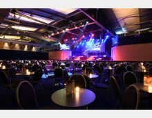 Großer Veranstaltungsraum mit Bühne, beleuchtet durch bunte Scheinwerfer und der Beschriftung "Stars in Concert". Im Vordergrund stehen zahlreiche runde Tische mit Kerzen und Bestuhlung, die für ein Publikum vorbereitet sind.