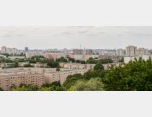 Stadtlandschaft mit großflächigen Wohnblöcken, überwiegend aus Beton, umgeben von Bäumen und Grünflächen; im Hintergrund sind weitere Gebäude, der Berliner Fernsehturm und eine flache, bewölkte Himmelsdecke zu sehen.