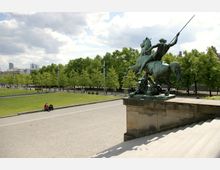 Bronzestatue eines Reiters mit erhobenem Speer auf einem Podest, im Vordergrund eine breite Steintreppe und eine gepflasterte Fläche, im Hintergrund eine Grünanlage mit Baumreihen und vereinzelt sitzenden oder spazierenden Personen.