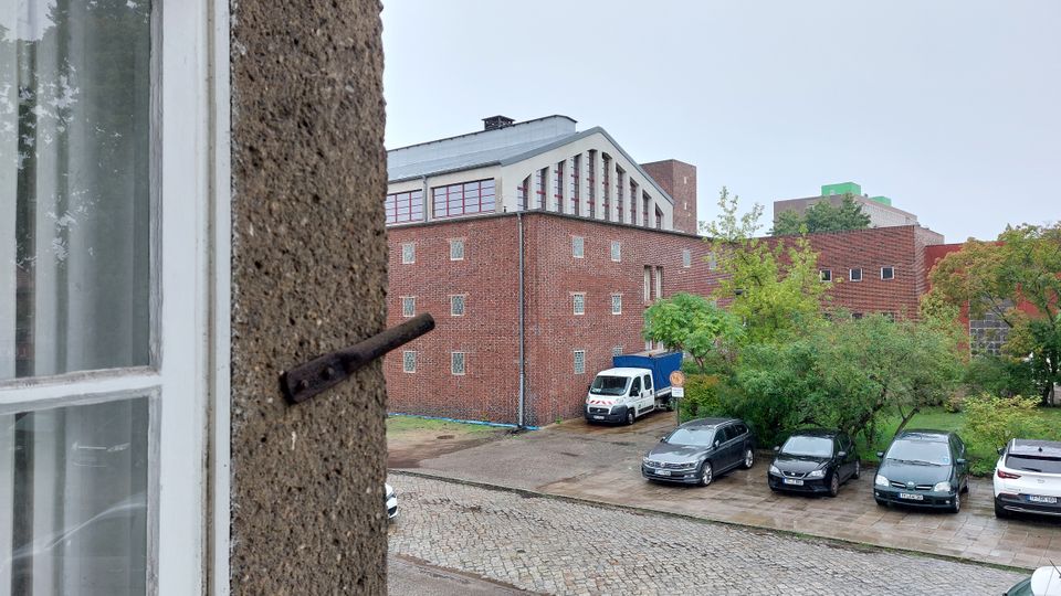 Blick aus einem Fenster auf ein modernes Backsteingebäude mit flachem Dach und großen Fenstern an der oberen Fassade, umgeben von Bäumen und Sträuchern. Im Vordergrund sind eine gepflasterte Zufahrt und mehrere geparkte Fahrzeuge zu sehen.