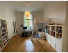 Ein helles Zimmer mit Holzboden und einer hohen Zimmerdecke, ausgestattet mit einem Hochbett, einem Arbeitsbereich mit Schreibtisch und Stuhl vor einem großen Fenster sowie mehreren weißen Regalen voller Bücher und Ordner. Im Hintergrund steht ein kleineres Sofa unter dem Hochbett, und eine moderne Deckenleuchte hängt zentral im Raum.