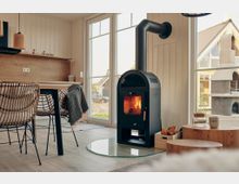 Gemütlicher Innenraum mit einem modernen schwarzen Kaminofen auf einer runden Glasplatte. Im Hintergrund große Fenster mit Blick auf eine ländliche Umgebung, daneben ein Essbereich mit Holztisch und geflochtenen Stühlen.